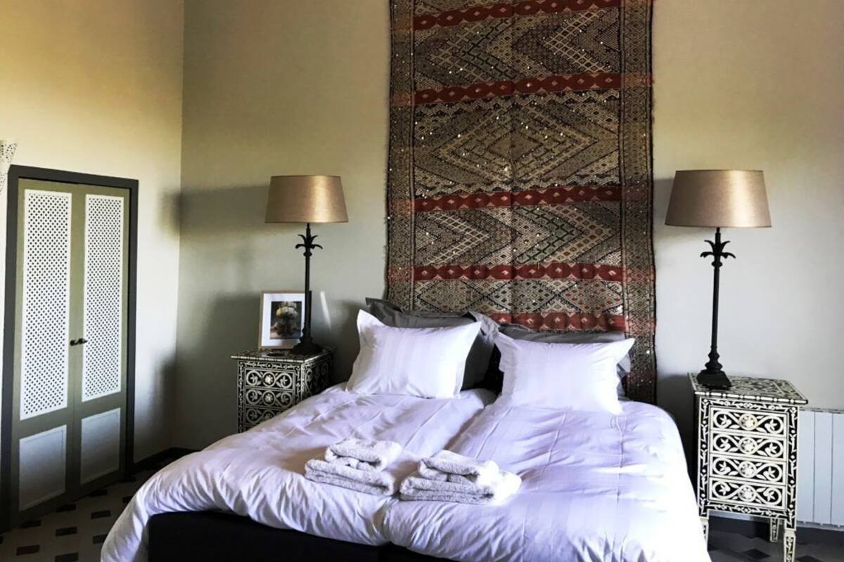 A neatly made bed with white linens and towels, two bedside tables with lamps, a patterned tapestry on the wall, and a door with a decorative grille.