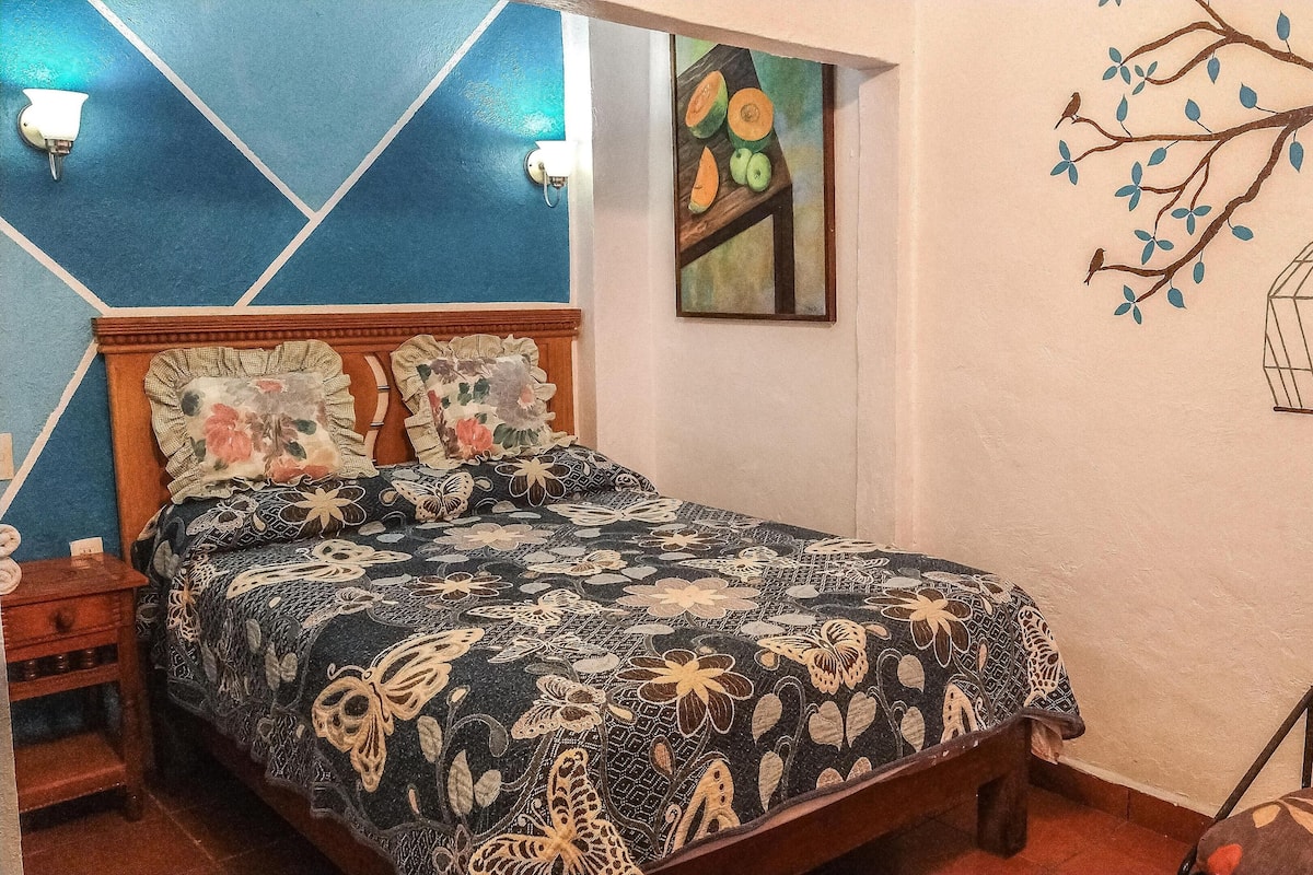 Een slaapkamer met een bed, nachtkastje, stoel en een muur met een wanddecoratie van een boom en een vogel.