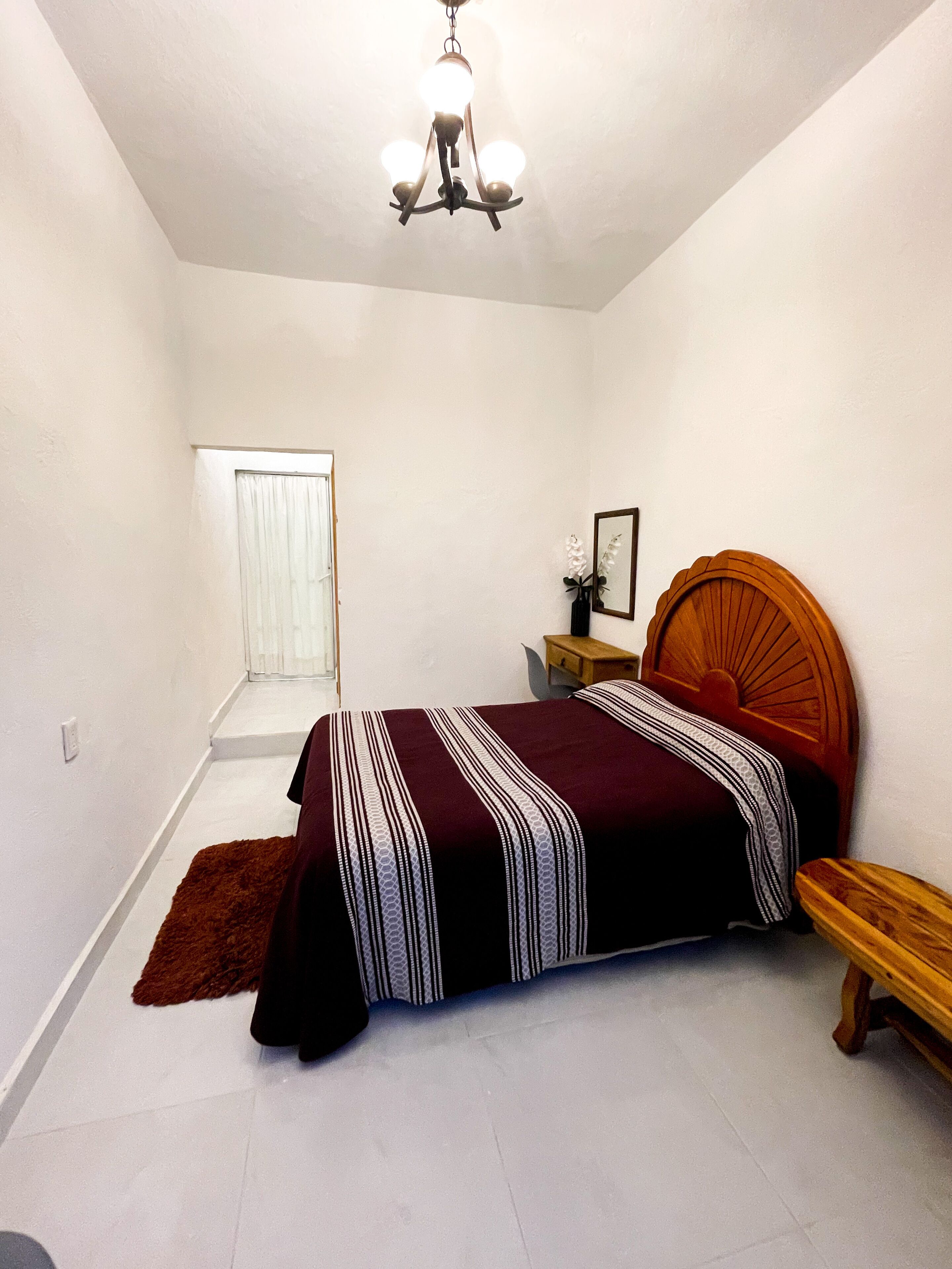 Une chambre à coucher comprenant un lit, une table de chevet en bois, un miroir et un luminaire fixé au plafond.