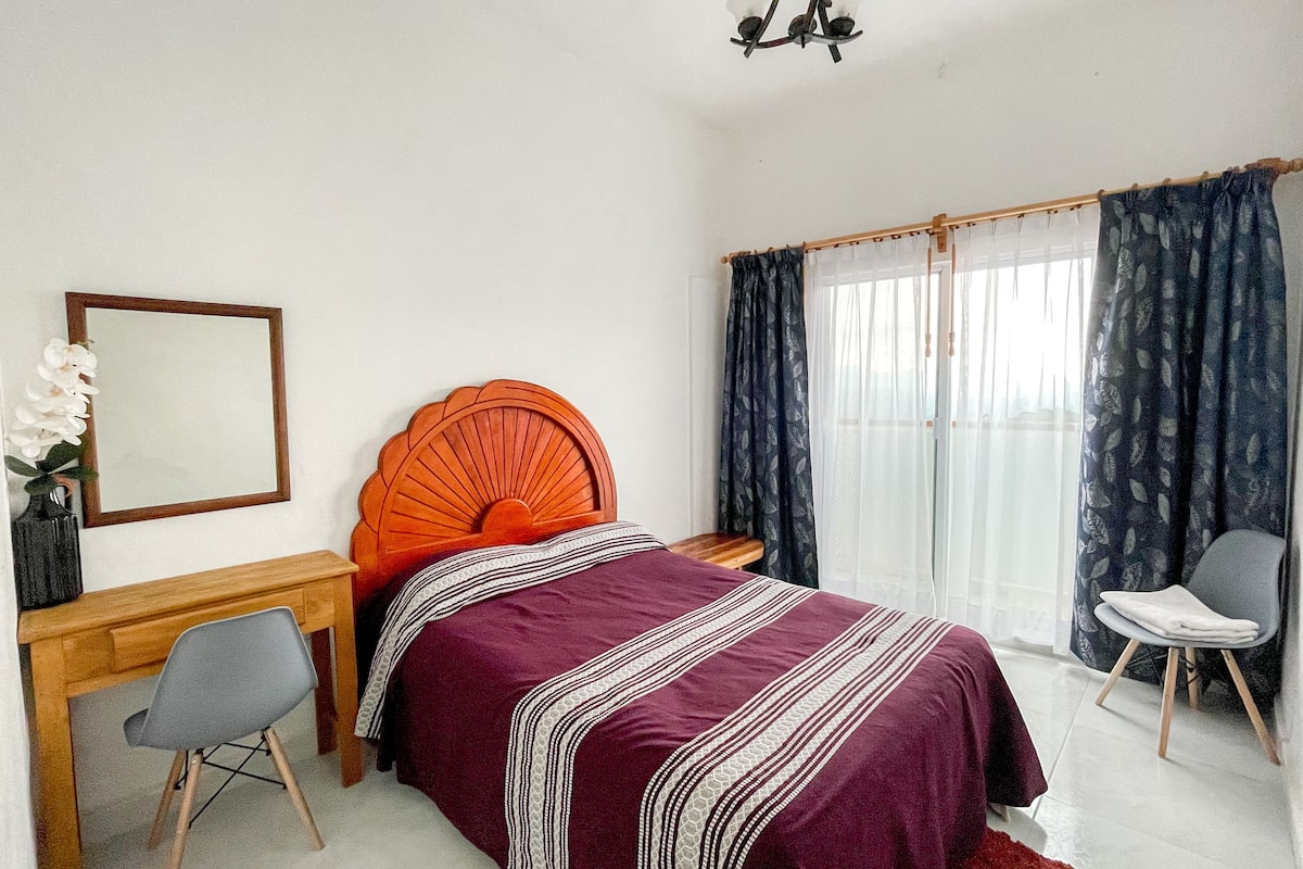 Een slaapkamer met een bed, een bureau, een stoel, een spiegel en een raam met gordijnen.