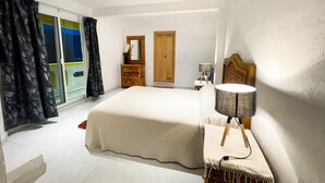 Une chambre à coucher avec un grand lit, une commode en bois, un miroir, une lampe et une porte donnant sur un balcon.
