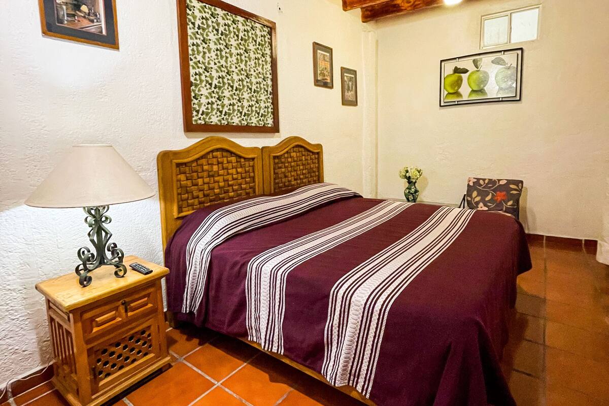 Een slaapkamer met een houten hoofdbord, een bed met een gestreepte dekbedovertrek, een nachtkastje met een lamp en een betegelde vloer.