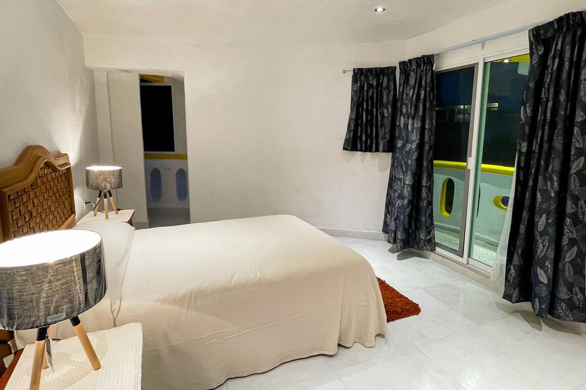 Een slaapkamer met een groot bed, een nachtkastje met een lamp, een hoofdbord en uitzicht op een balkon door een glazen deur.