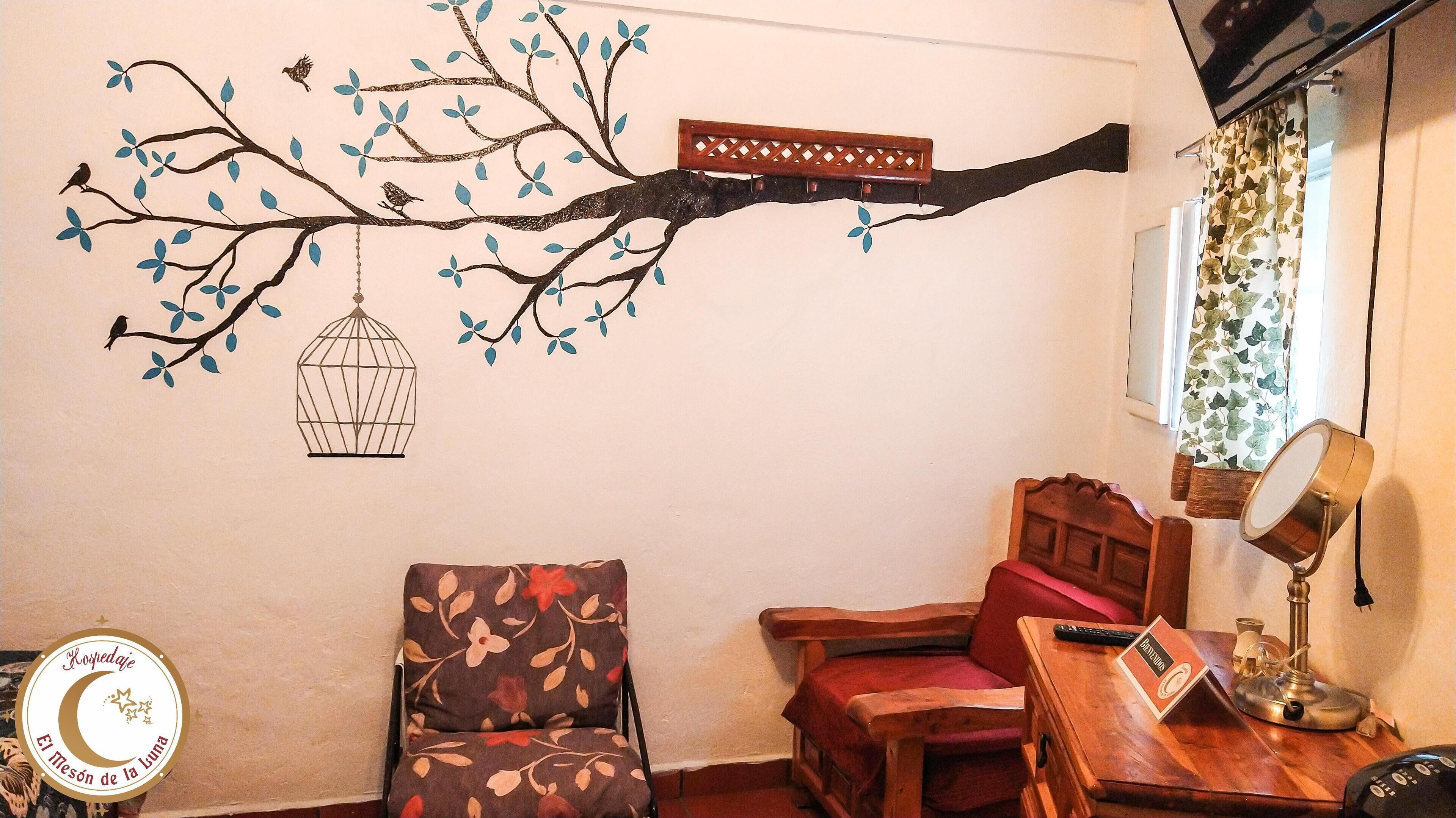 Quarto com um mural de árvore, pássaros e uma gaiola suspensa. Há uma poltrona com padrão floral, uma cadeira de madeira com almofada vermelha e uma pequena mesa de madeira com uma luminária e um controle remoto.