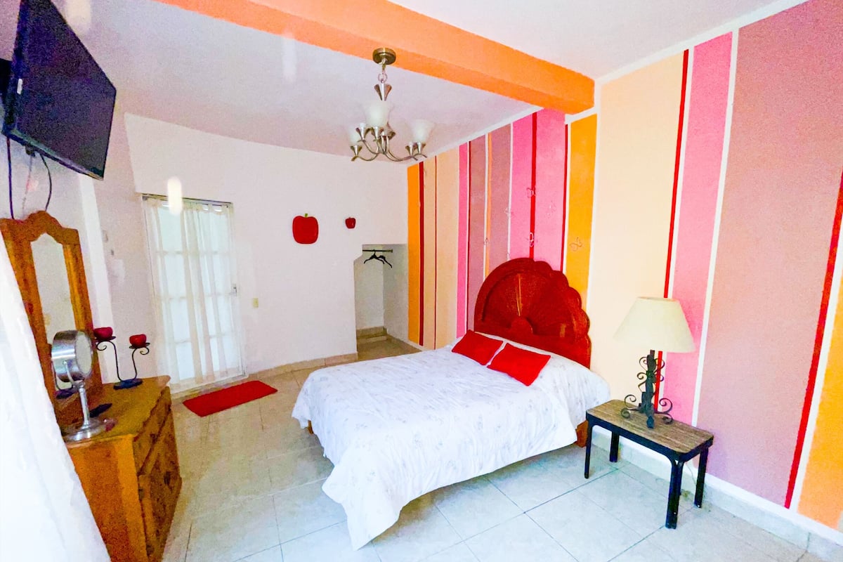 Een slaapkamer met een bed, nachtkastje met lamp, dressoir, spiegel en televisie.