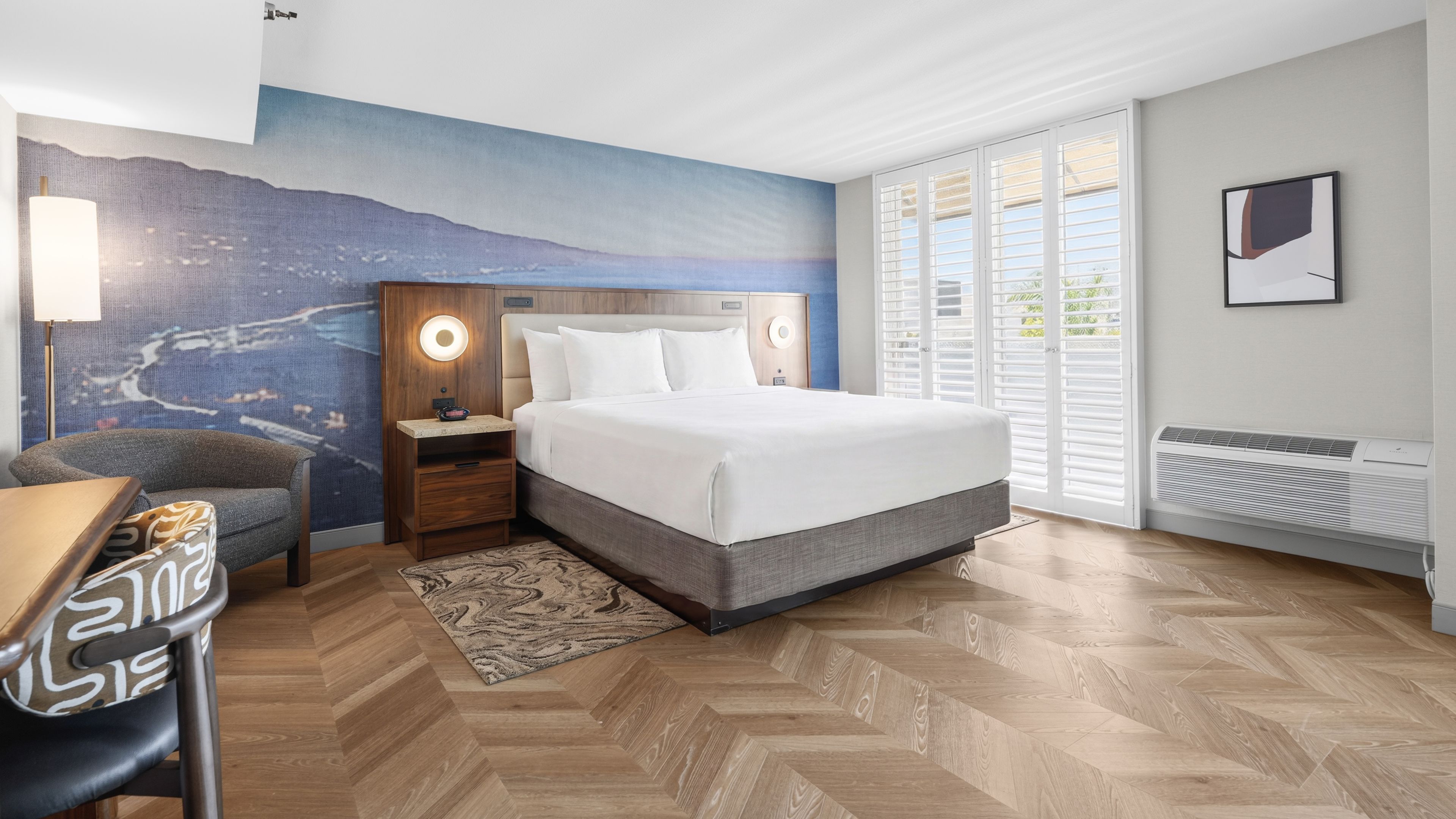 A hotel room with a bed, a nightstand, a desk, a chair, a large window with blinds, and a wall mural.