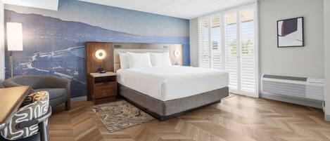 A hotel room with a bed, a nightstand, a desk, a chair, a large window with blinds, and a wall mural.