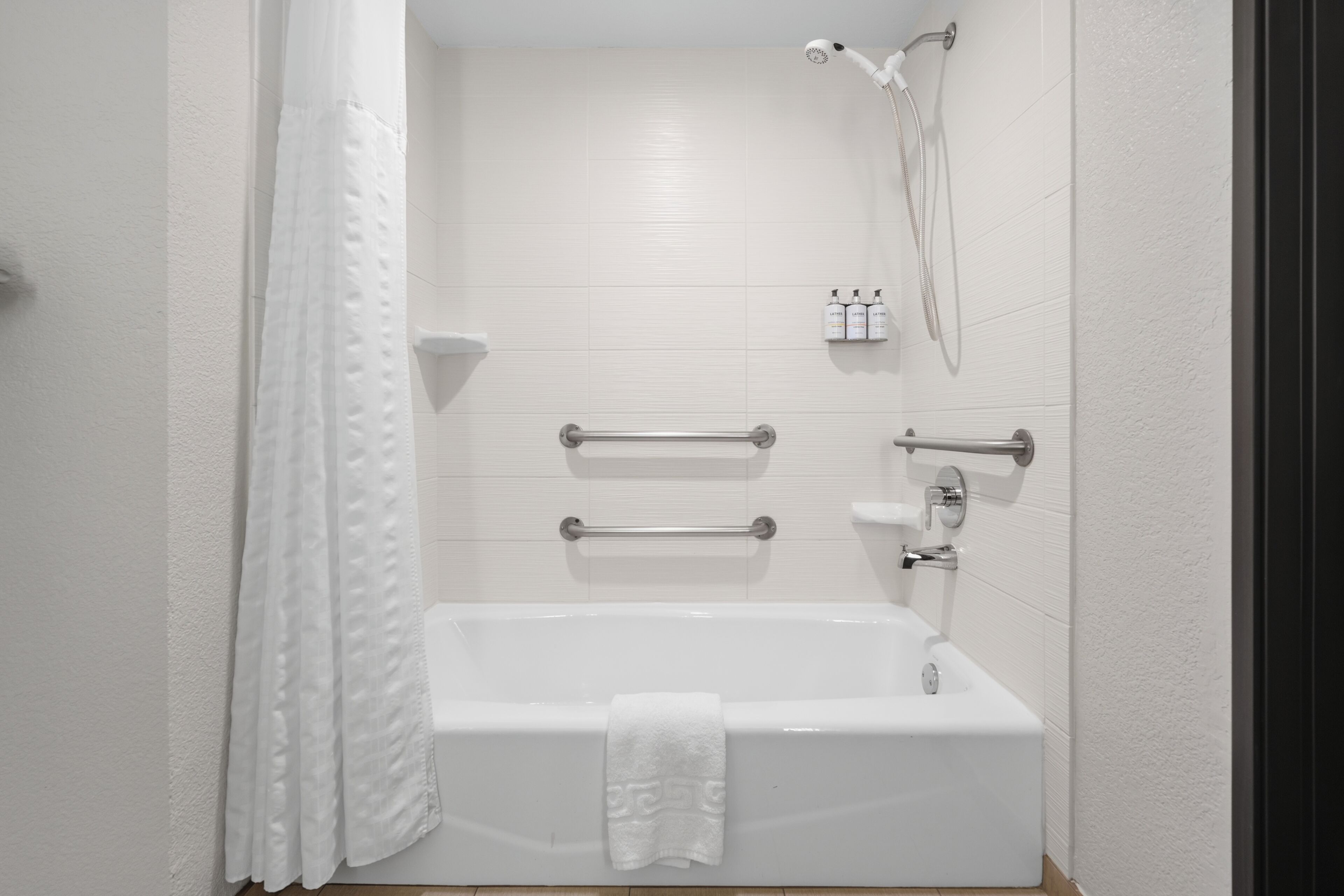 A modern bathroom with a white bathtub, grab bars, a showerhead, and toiletries on a shelf.