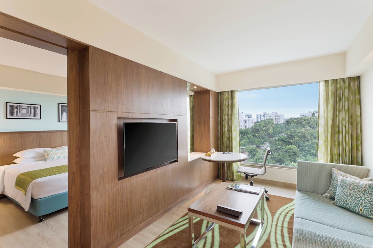 A hotel room with a bed, a sofa, a TV, a small table, and a window with a view of buildings and trees.