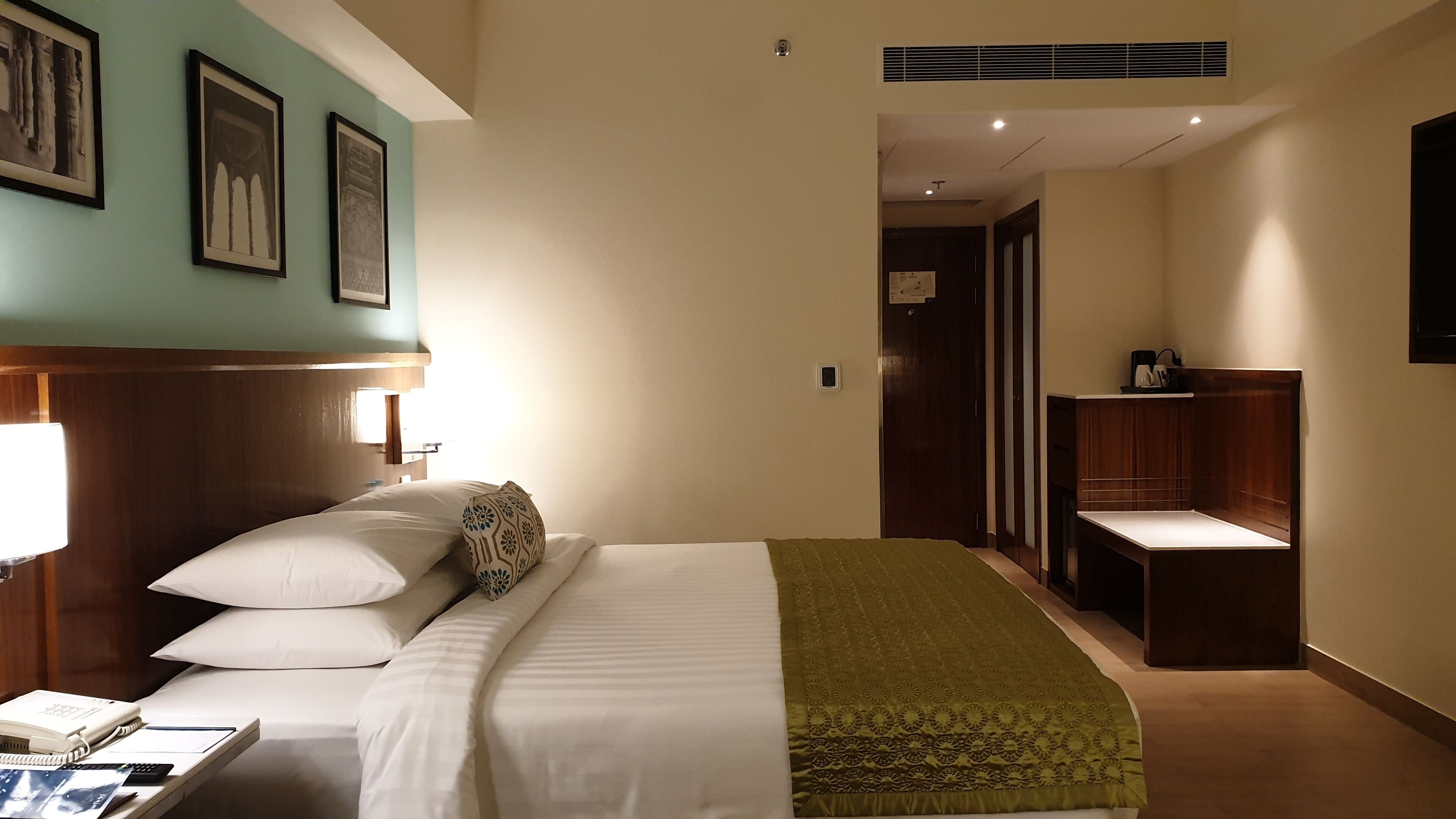 A hotel room with a bed, bedside table, lamp, and a desk with a phone and papers.