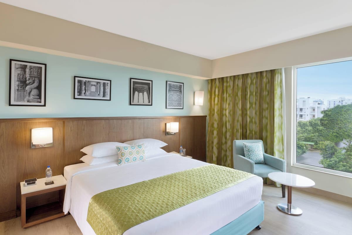 A hotel room with a large bed, a nightstand, a chair, a small table, and a window with a view of buildings and trees.