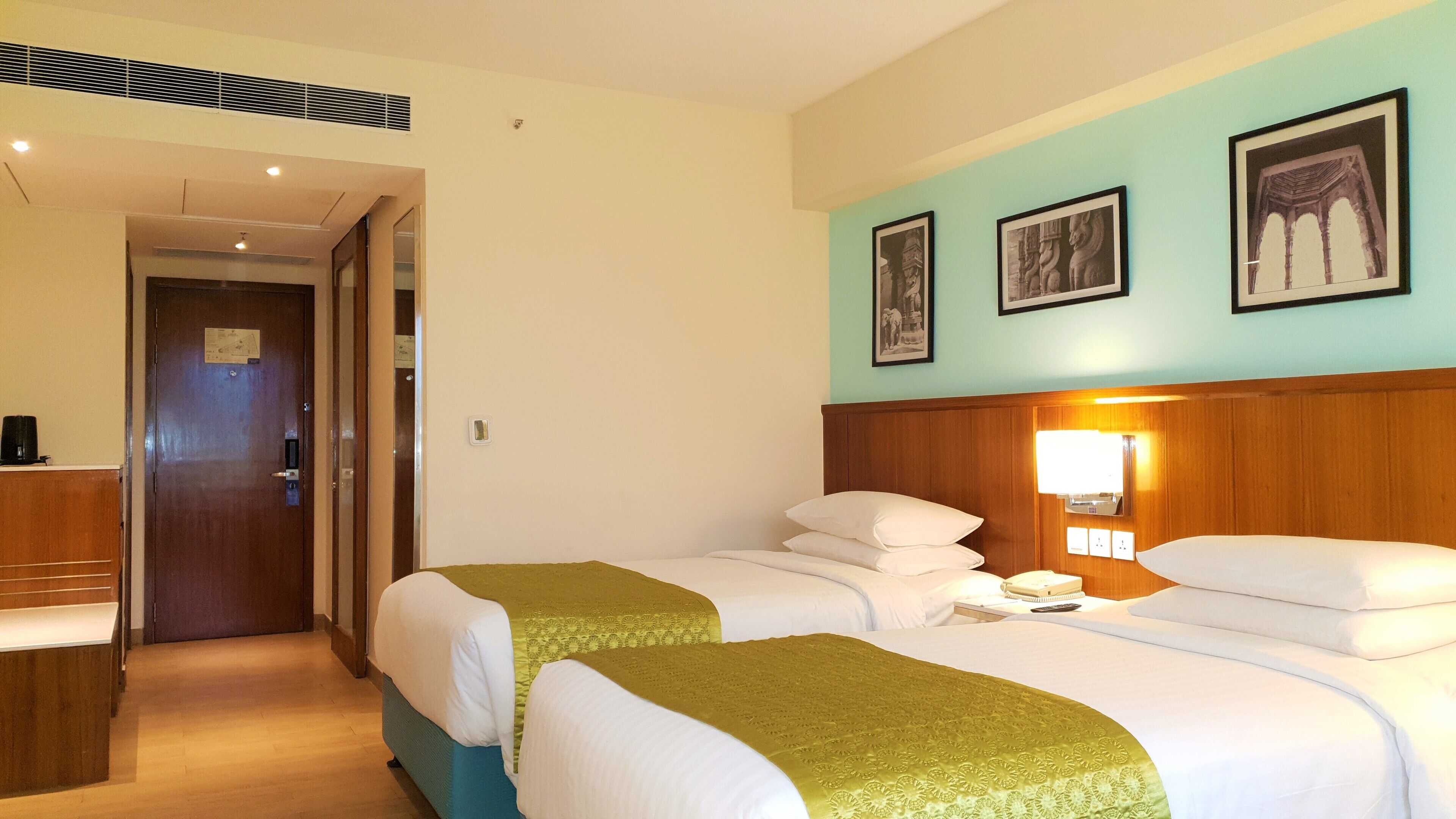 A hotel room with two beds, a wooden headboard, a nightstand, a desk, and a door leading to a hallway.