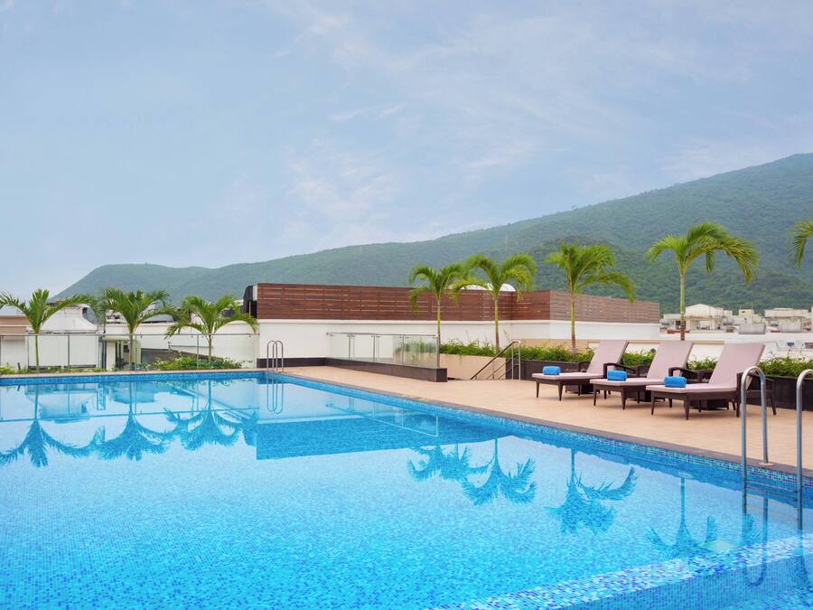 Una piscina con sillas de descanso y palmeras, rodeada de edificios y una montaña al fondo.