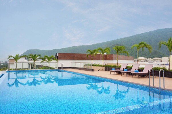 A swimming pool with lounge chairs and palm trees, surrounded by buildings and a mountain in the background. - Fairfield by Marriott Visakapatnam (Visakhapatnam)