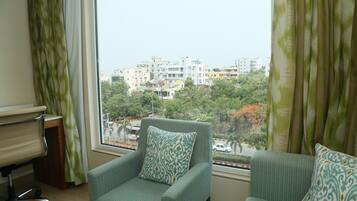 A room with a view of a cityscape through a window, a green armchair with a patterned pillow, and a small desk with a chair.