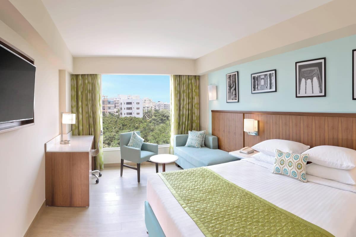 A hotel room with a bed, a desk, a chair, a small table, and a view of the city.