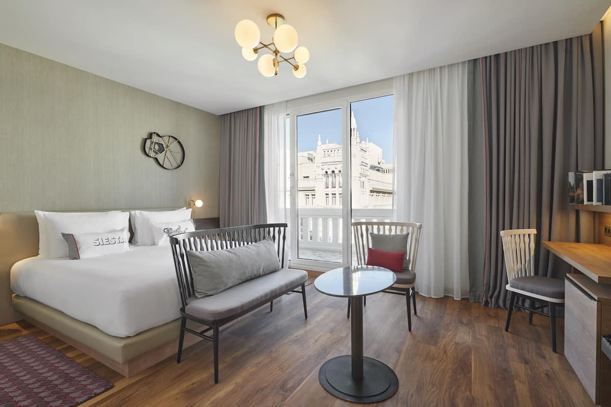 Una camera d'hotel con un letto, un divano, un tavolino rotondo e una sedia. Si vede un edificio dalla finestra.