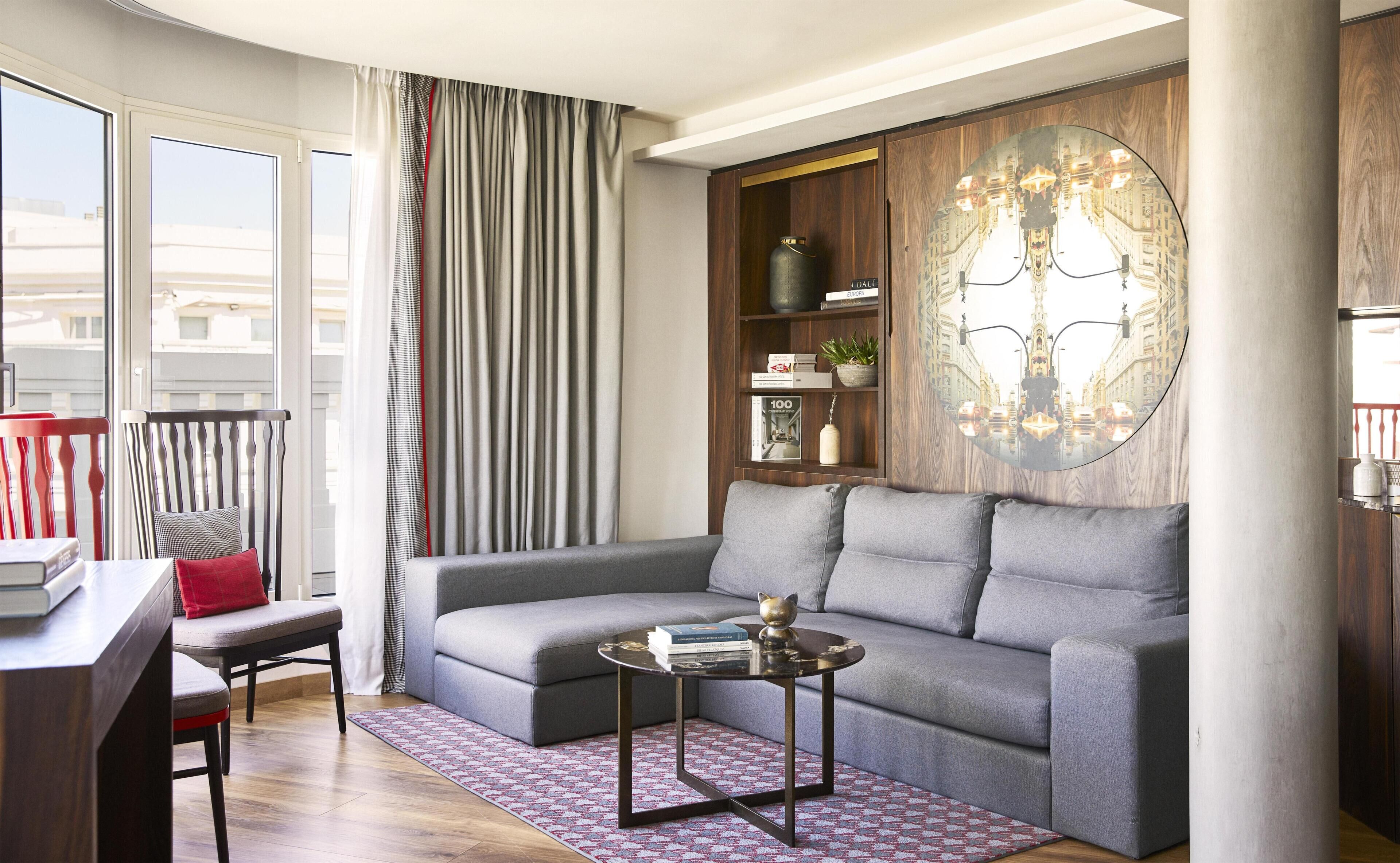 Una sala de estar moderna con un sofá gris, una mesita redonda, una silla roja y una estantería con libros y objetos decorativos.