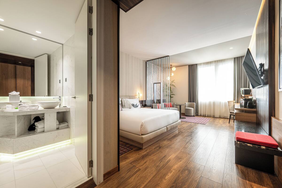 Una moderna camera d'albergo con un letto grande, una zona salotto separata, un bagno con un vanitino e uno specchio ampio.