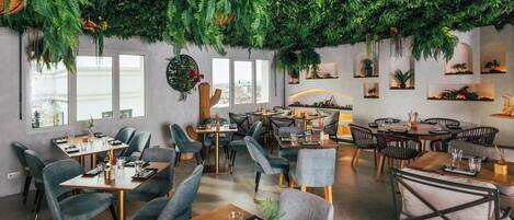 Un restaurante con varias mesas y sillas, un techo con plantas colgantes, y vistas a la ciudad a través de grandes ventanales.
