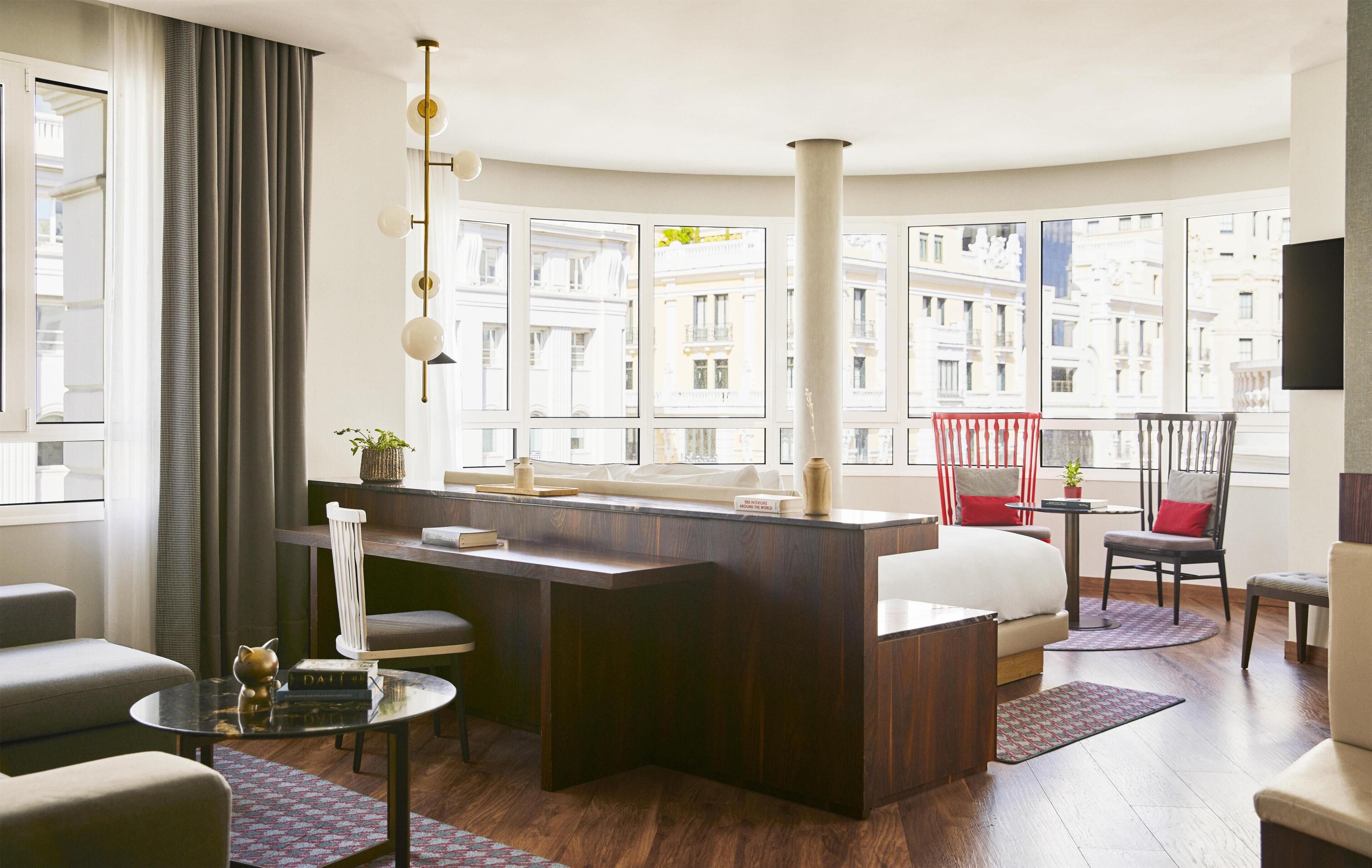 Una habitación de hotel moderna con un escritorio de madera, un sofá, una silla, una mesita y un amplio ventanal con vistas a edificios.