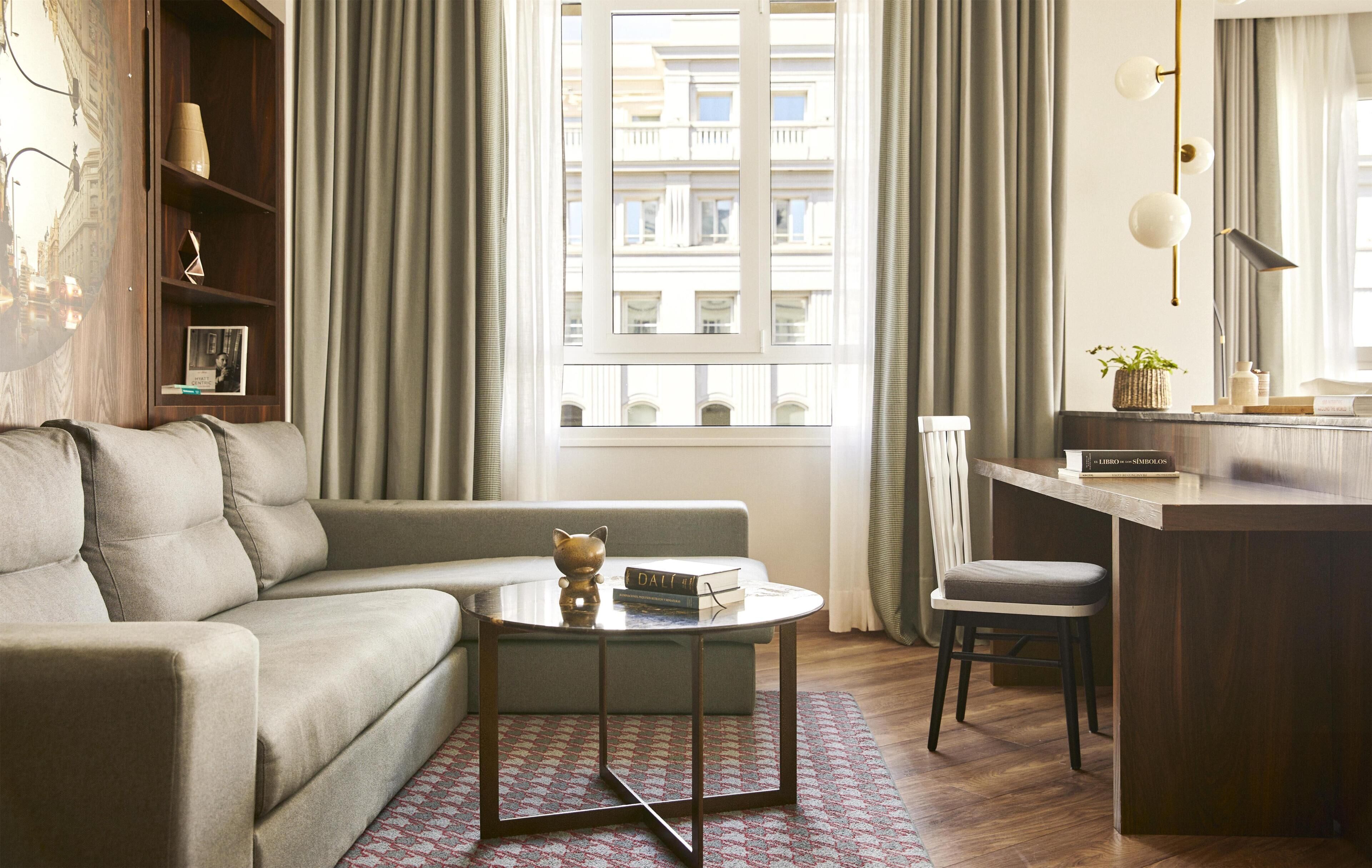 Una sala de estar moderna con un sofá, una mesa de centro redonda, un taburete pequeño, una estantería y vistas a edificios a través de una ventana.