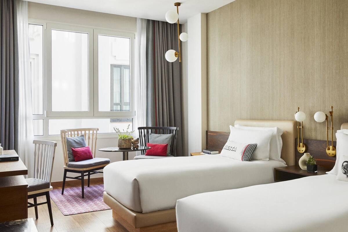 Camera d'albergo con due letti, una scrivania, una sedia, un tavolino e una finestra con tende.