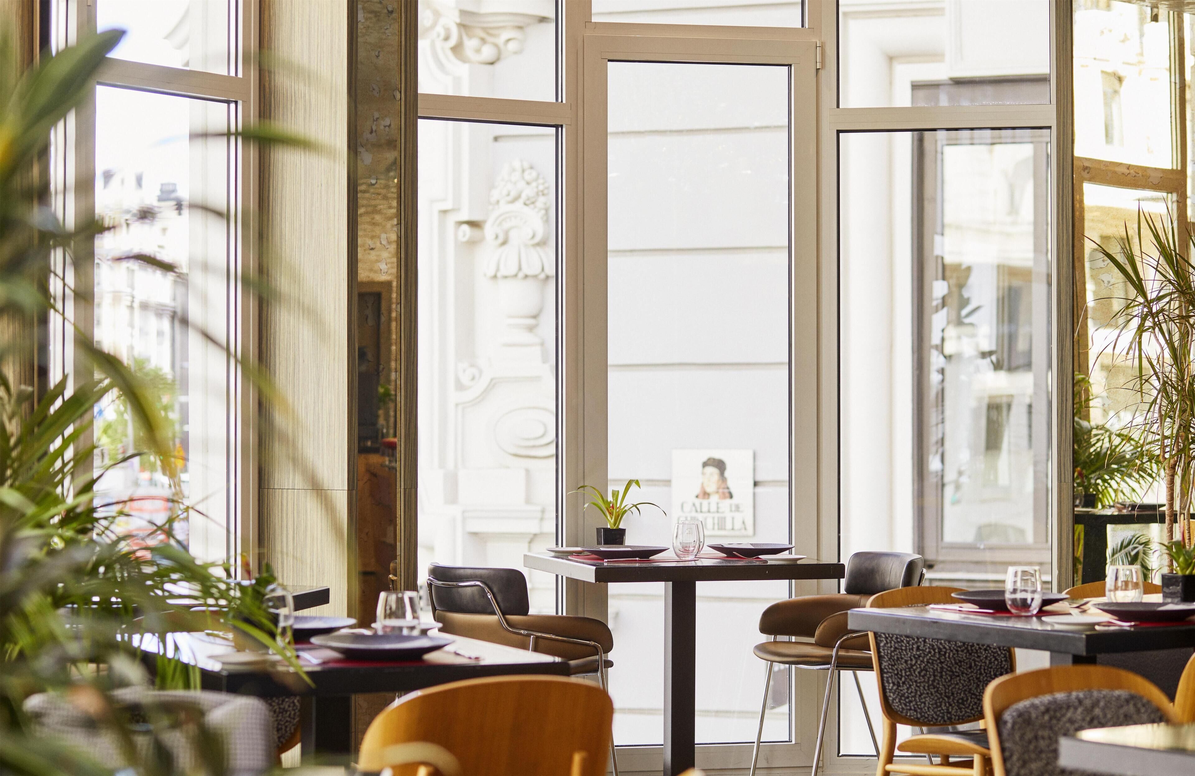 Een restaurantinterieur met grote ramen, tafels, stoelen en potplanten.