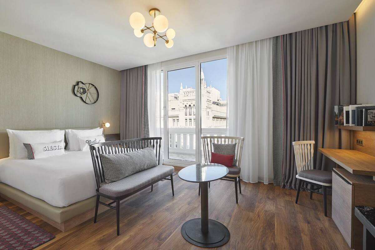 Una camera d'albergo con un letto, un tavolo rotondo, un divano, una scrivania e vista sui palazzi attraverso ampie finestre.