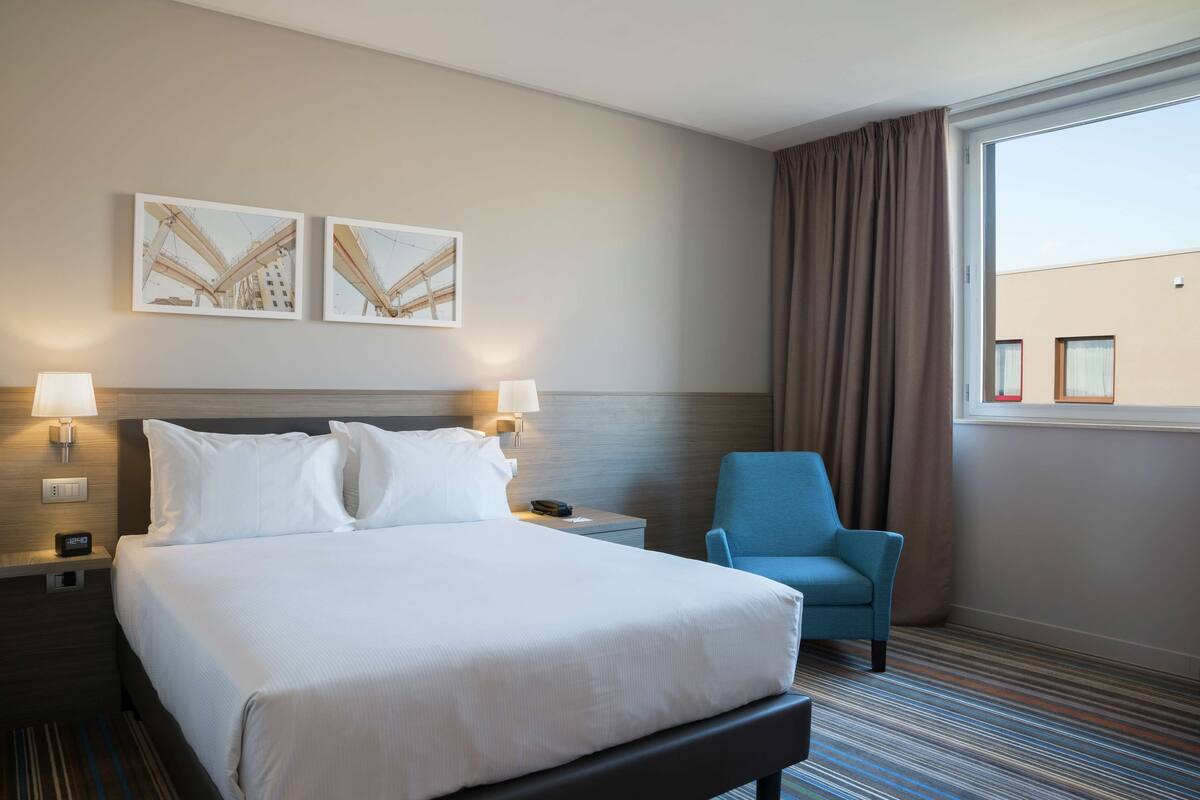 A hotel room with a large bed, two wall-mounted lamps, a blue armchair, and a window with curtains.