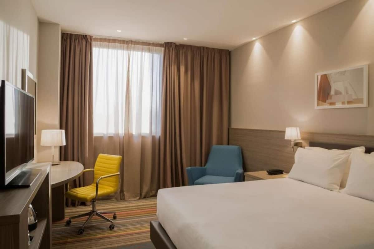 A hotel room with a bed, a desk with a chair, a television, and a window with curtains.