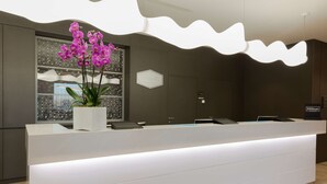 A modern hotel reception area with a white reception desk, a potted orchid, and a digital display. - Hampton by Hilton Rome East (Rome)