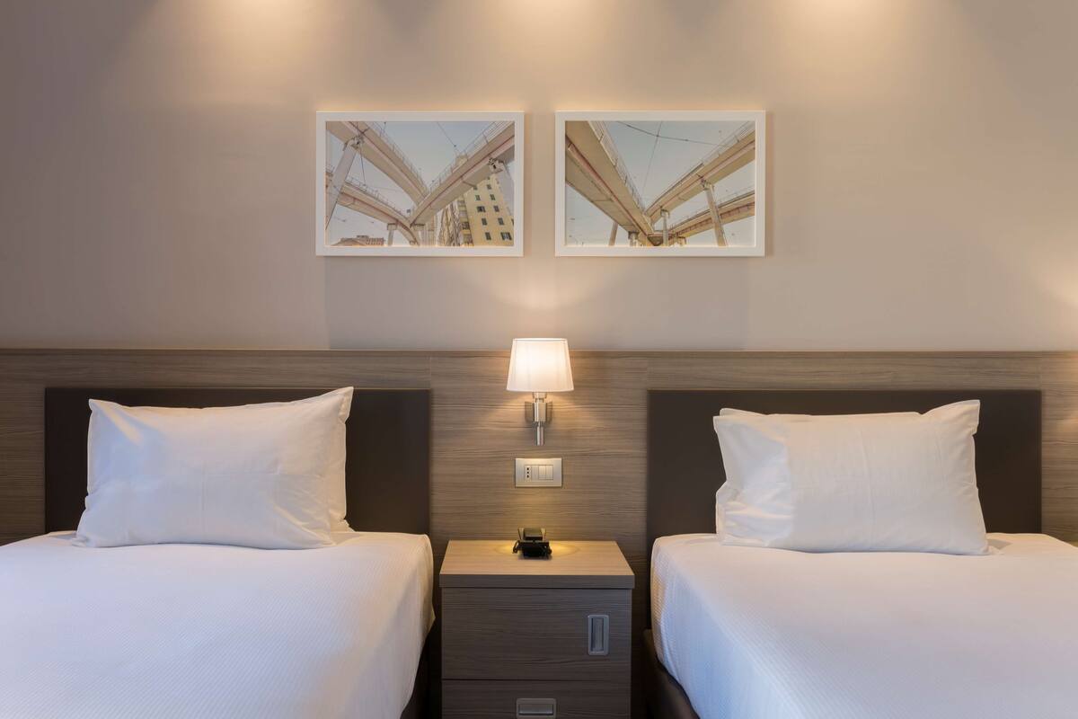 A hotel room with two beds, a nightstand with a lamp, and two framed pictures on the wall.