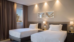 A hotel room with two beds, a window with curtains, wall-mounted lamps, and framed pictures. - Hampton by Hilton Rome East (Rome)