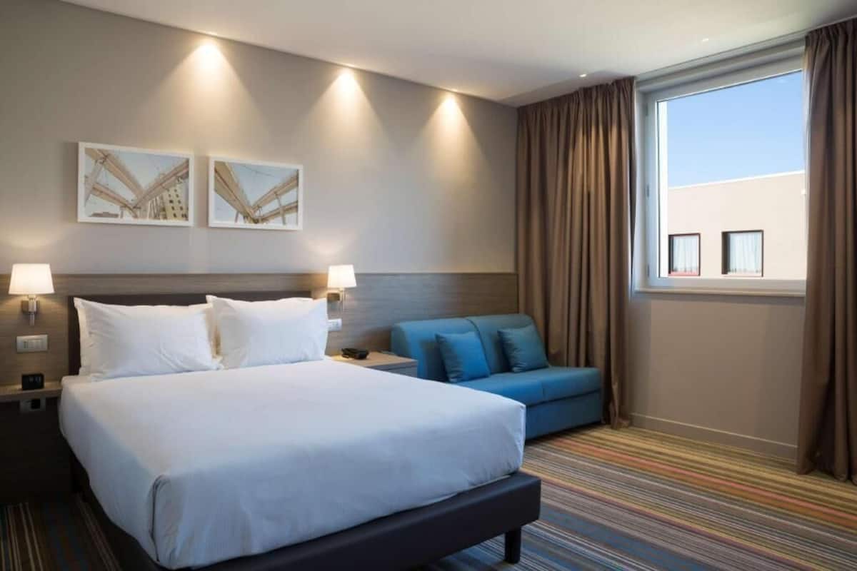 A hotel room with a large bed, a sofa, two wall-mounted lamps, and a window with curtains.