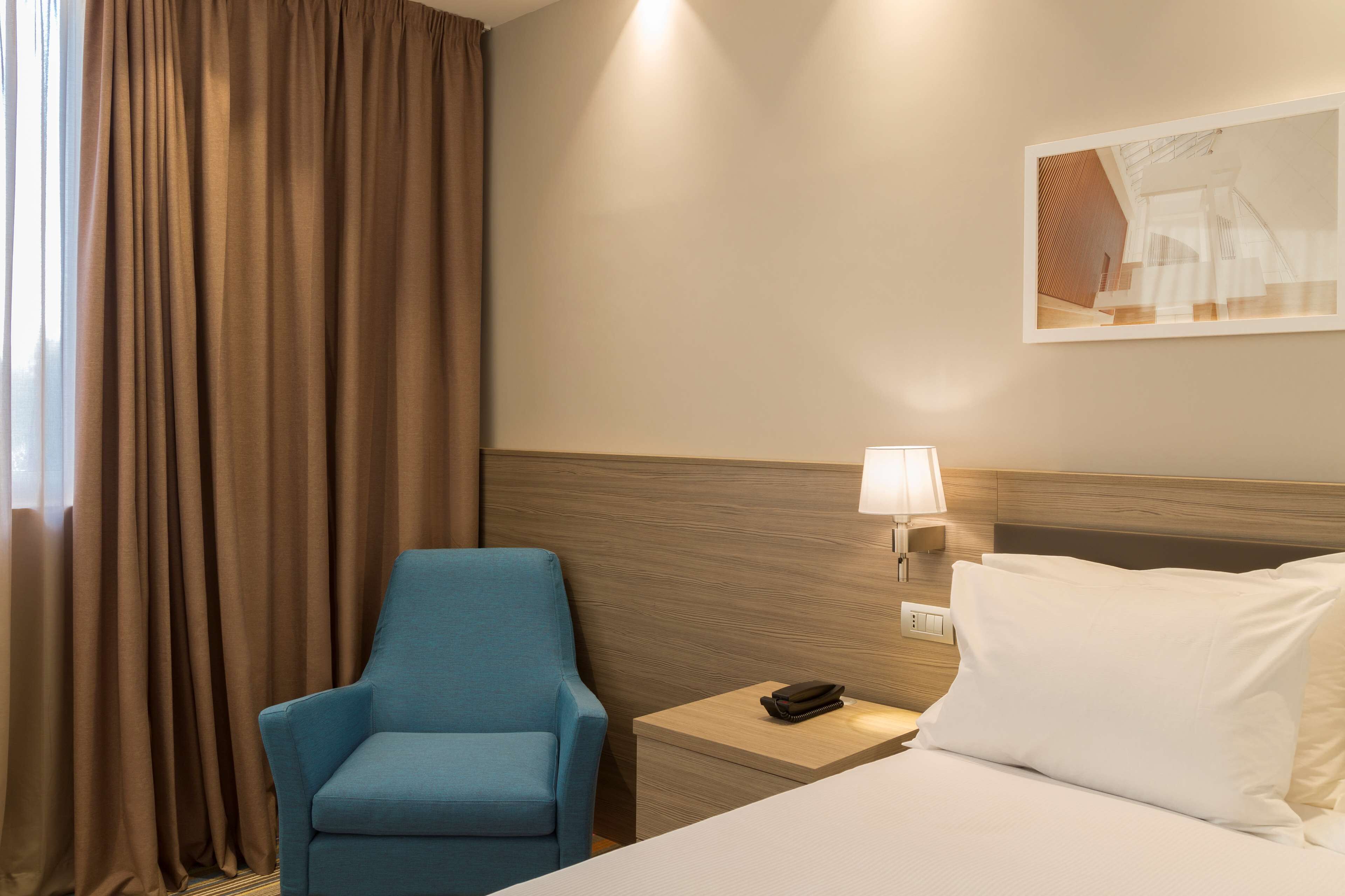 A hotel room with a bed, a blue armchair, a nightstand with a lamp, and a wall-mounted artwork.