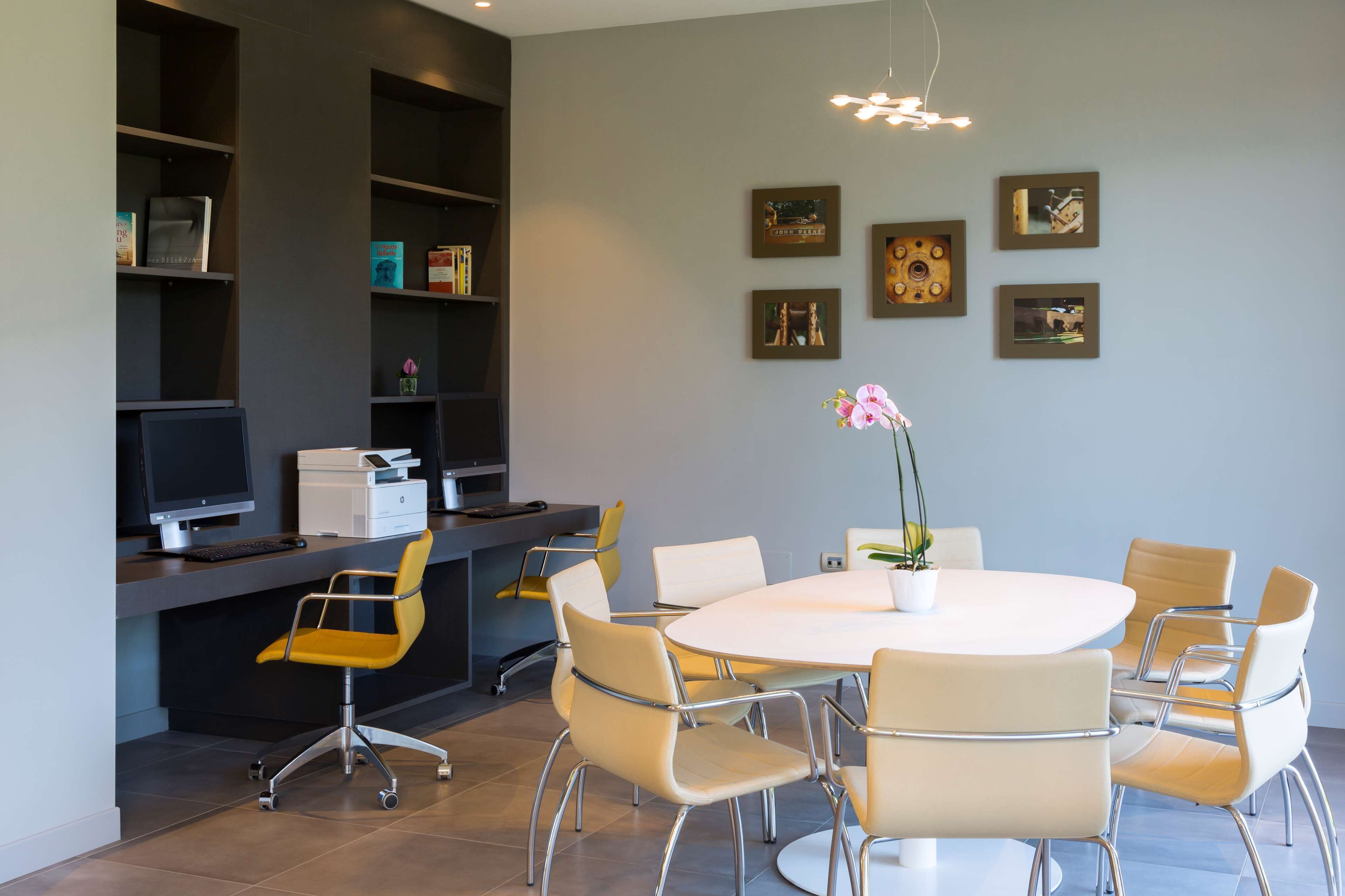 A modern meeting room with a round table, six chairs, a computer, and shelves with books and decorative items.