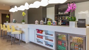 A hotel reception area with a white counter, bar stools, a minibar, and a minibar refrigerator stocked with beverages. - Hampton by Hilton Rome East (Rome)