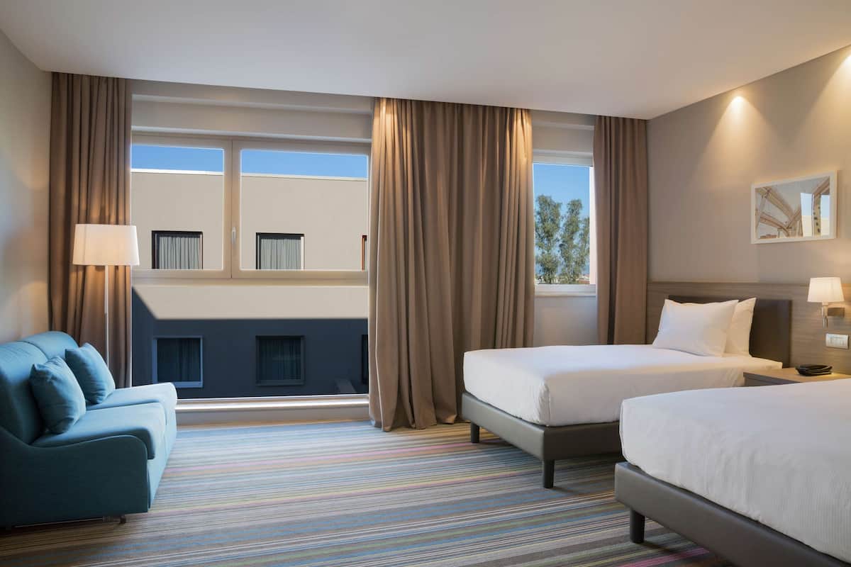 A hotel room with two beds, a sofa, a window with curtains, and a striped carpet.