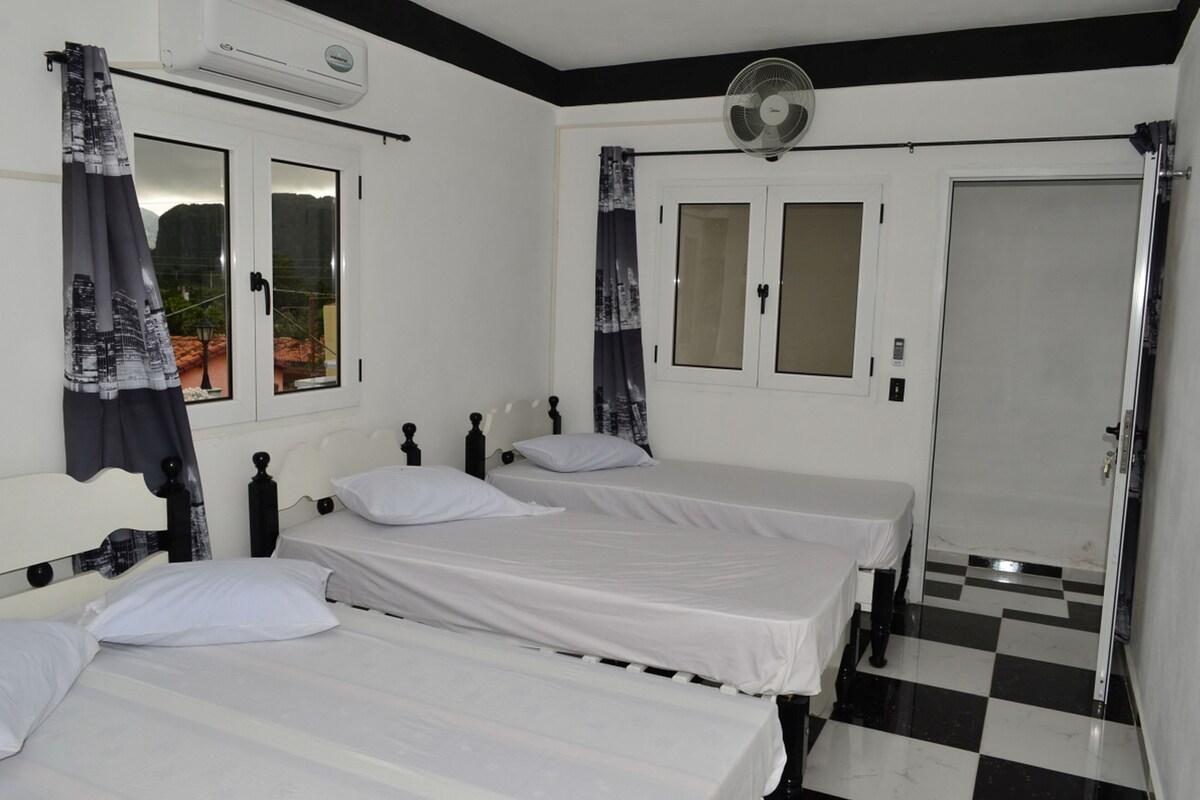 Ein Zimmer mit drei Einzelbetten, einem Deckenventilator, einer Klimaanlage und einem Schachbrettboden.