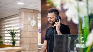 A man at a reception desk in a hotel lobby with a wooden accent wall and a potted plant. - Aviv Holiday Flat (Tiberias)