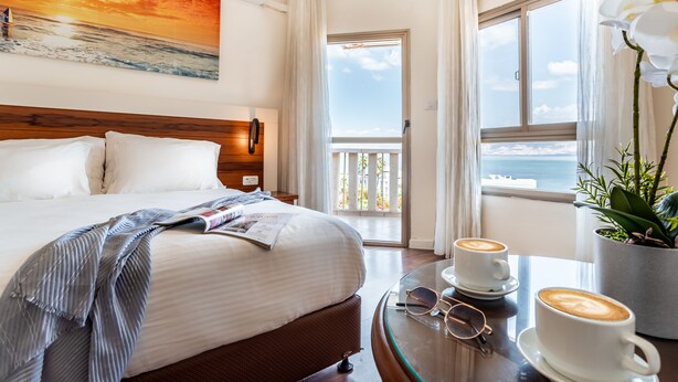 A hotel room with a bed, a table with two cups of coffee, a pair of glasses, and a window with a view of the sea. - Aviv Holiday Flat (Tiberias)
