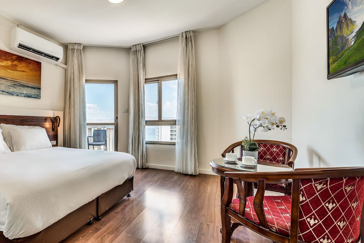 A hotel room with a bed, bedside table, a chair, a small table with a plant, and a painting of a sunset.