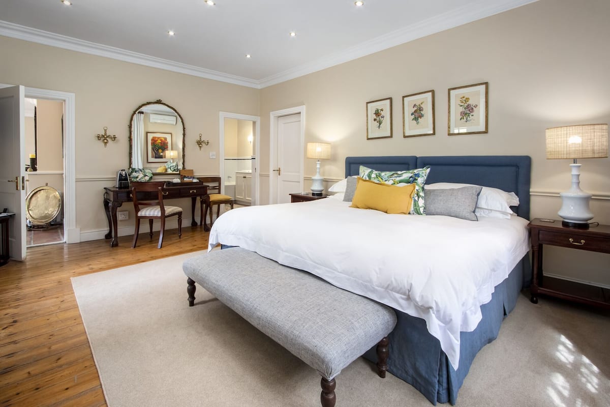 Un dormitorio con una cama grande, un banco, un escritorio con silla, un espejo y cuadros con motivos florales en la pared.