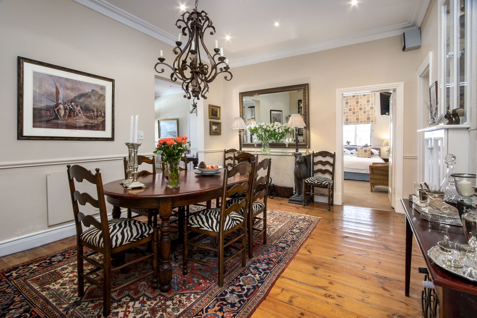 A dining area with a wooden table, chairs, and a chandelier. There is a painting on the wall and a mirror above the table.