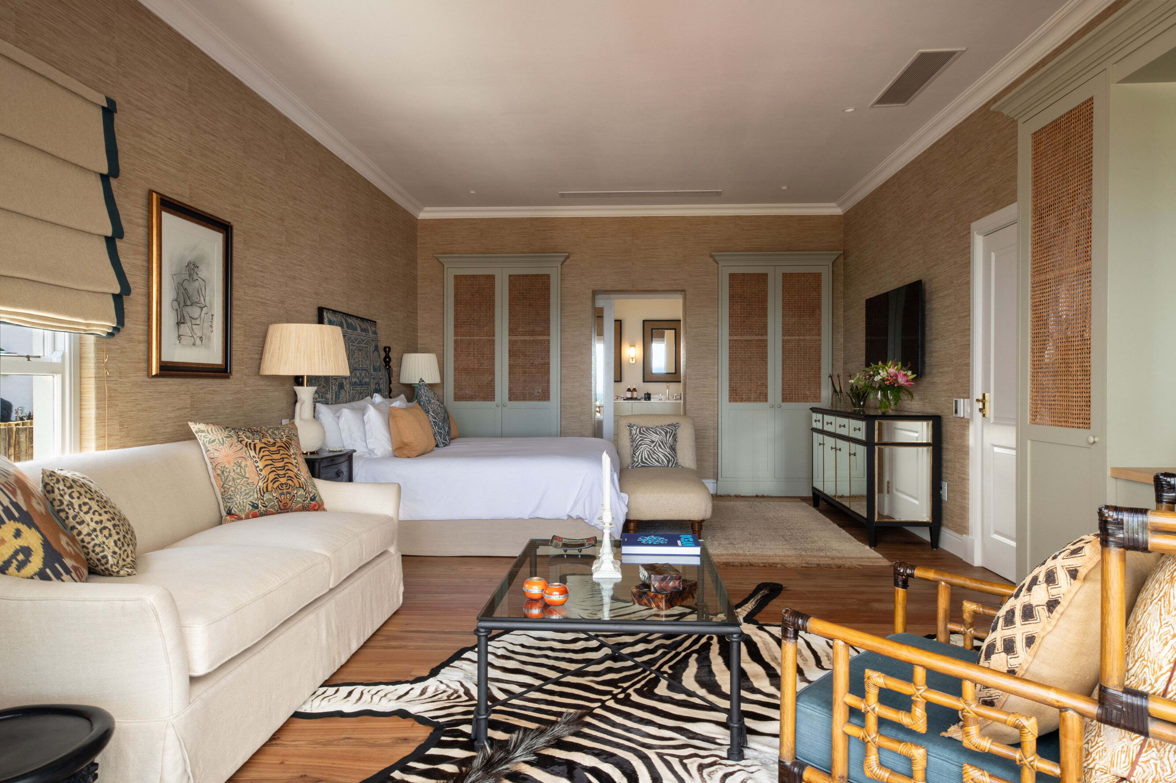 A spacious hotel room with a large bed, a sitting area with a sofa and armchairs, a coffee table, and a view of the hallway.