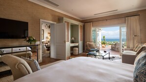 Une chambre d’hôtel spacieuse avec un grand lit, un coin salon offrant une vue sur la mer, une télévision et un balcon avec des sièges d’extérieur.