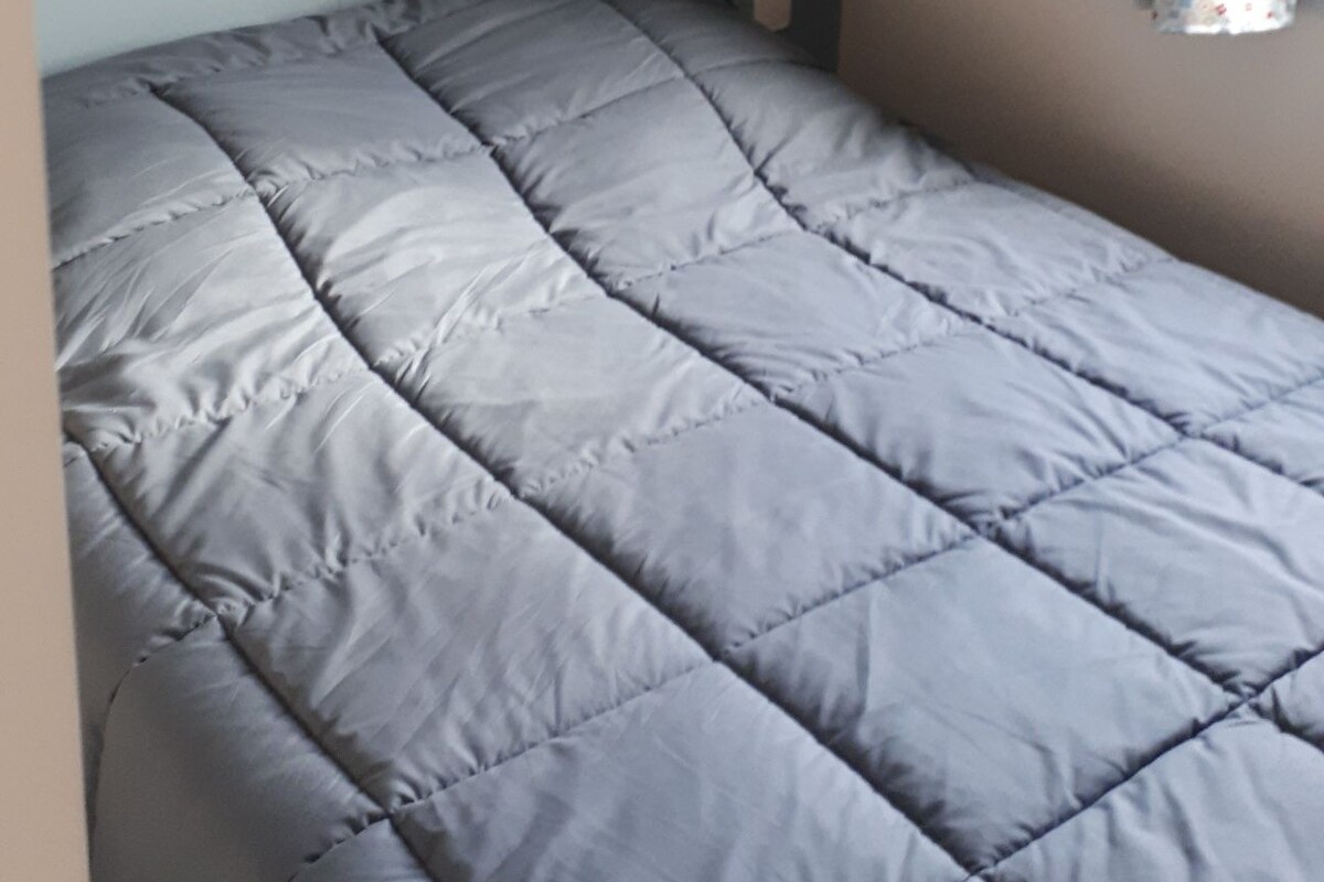 A single bed with a quilted grey cover, a wooden shelf above it, and a small bedside table with a lamp.