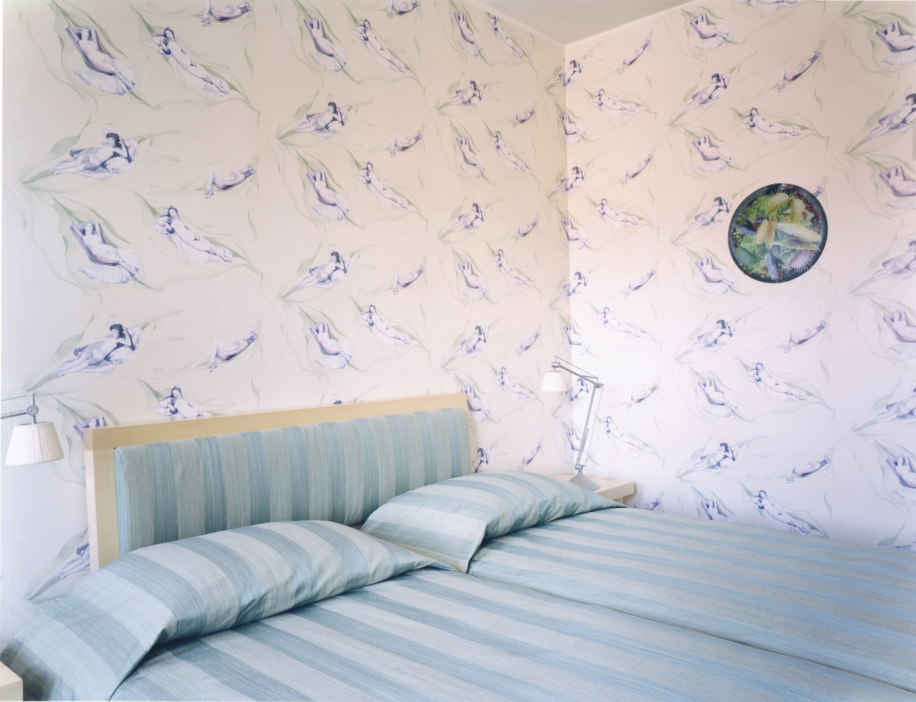 A bedroom with a bed, striped bedding, a headboard, a nightstand, a lamp, and a wall with a repeating pattern.