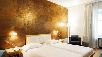 A hotel room with two beds, a wooden headboard, a nightstand with a lamp, a chair, and a view of the outdoors.